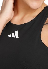 Close-up van een persoon in een zwarte mouwloze top met een wit Adidas-logo, hand omhoog bij het oor, met een tatoeage op de schouder.