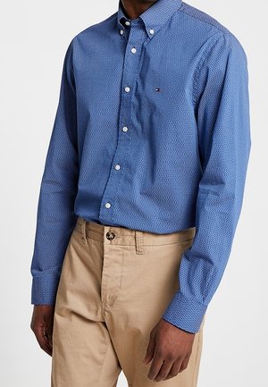 Mann trägt ein blau gemustertes Hemd mit Knöpfen, in beige Hosen gesteckt, steht vor einem schlichten Hintergrund mit entspannten Händen.