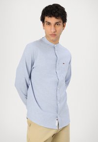 Camicia azzurra a maniche lunghe con colletto alla cinese, bottoni frontali e una piccola patch con il logo sulla tasca del petto. Tessuto morbido e texturizzato.