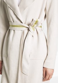 Beige mockakappa med sjalkrage, bältesmidja med ett gult-strimmigt bälte och två framfickor med synliga sömsdetaljer.