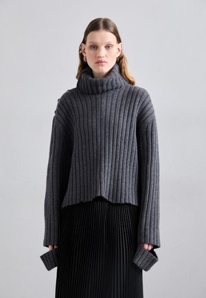 Szürke bordázott garbós pulóver, rövidített aljjal és hosszú ujjal, kiterjesztett mandzsettával. Egy fekete plisszírozott szoknyával kombinálva.