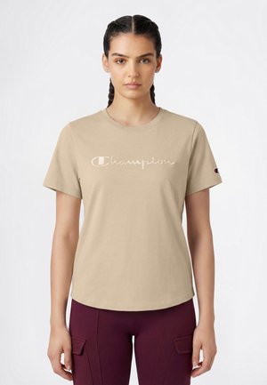 Junge Frau mit geflochtenem Haar trägt ein beiges Champion-T-Shirt und dunkelviolette Hose, steht vor einem einfarbigen weißen Hintergrund.