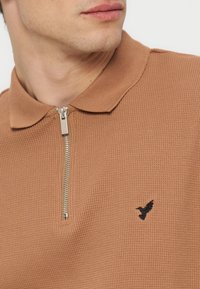 Κοντινό πλάνο ενός άνδρα που φοράει καφέ ραμμώδη πόλο με φερμουάρ και έμβλημα μικρού μαύρου πουλιού στο αριστερό στήθος.