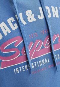 Sudadera azul con tejido texturizado, que presenta un gran logo blanco y rosa que dice "JACK & JONES SUPER" con un cuello de cordón.