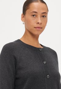 Grå cardigan med rund halsringning, knappstängning fram och ribbad manschett. Tillverkad av mjukt tyg med en slät yta och en lätt glans.