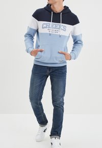 Sudadera con capucha en azul y azul marino con rayas blancas y azules claras, que cuenta con un bolsillo frontal y un diseño impreso que dice "CREEKS ORIGINAL".