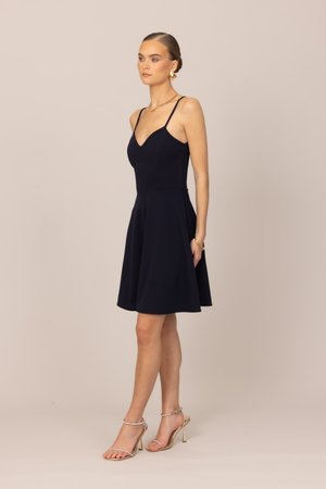 Kvinne i en svart ermeløs kjole med tynne stropper, gylne øredobber og armbånd, og gylne sandaler med høye hæler, stående foran en ensfarget bakgrunn.