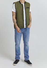 Chaleco acolchado de color verde oliva sin mangas con un bolsillo con cremallera negra, llevado sobre una camiseta blanca y combinado con unos jeans azul claro y zapatos negros.