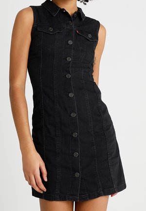 Czarna, bezrękawnikowa sukienka z denimu z guzikami z przodu i kołnierzykiem, noszona przez osobę z tatuażem na lewej ręce.