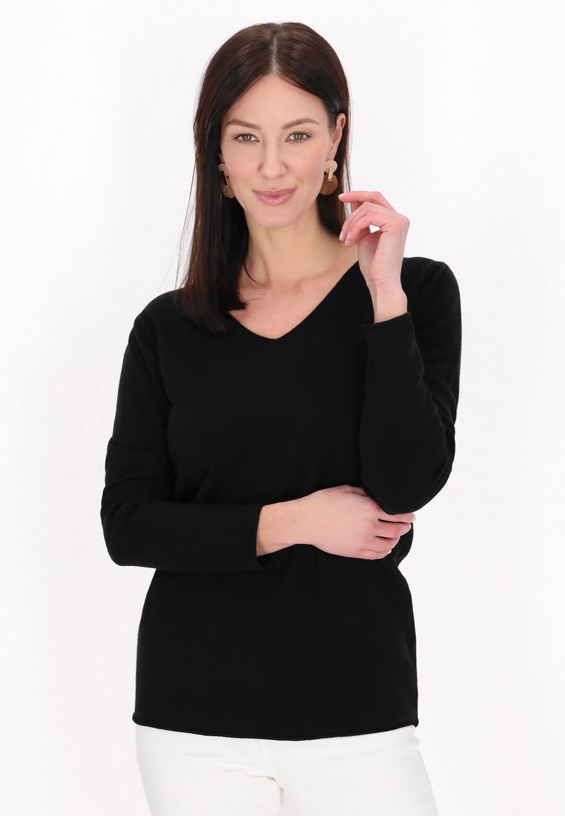 Pulover negru cu decolteu în V, mâneci lungi, țesătură moale, croială lejeră. Modelul pozează cu o mână pe braț, subliniind simplitatea și designul articolelor.
