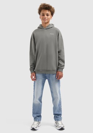 Tienerjongen met krullend haar staat in een grijze hoodie, lichtblauwe jeans en witte sneakers tegen een effen achtergrond.