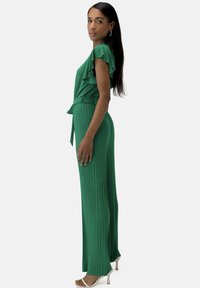 Mono verde plisado con mangas rizadas, detalle de cintura ajustada y diseño ceñido. Tela suave, silueta elegante y longitud hasta el tobillo.