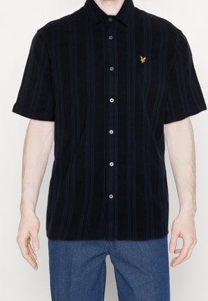 Man draagt een zwart overhemd met korte mouwen met verticale strepen en een klein geel vogel-logo op de borst, gecombineerd met een blauwe broek.