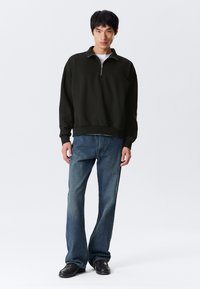 Jeune homme portant un sweat à col zippé noir, un t-shirt blanc, un jeans bleu et des chaussures noires, se tenant contre un fond clair uniforme.