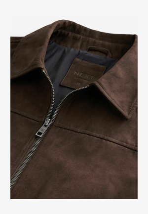 Veste en faux suède marron avec fermeture éclair, dotée d'un col et d'une étiquette de marque à l'intérieur. Texture lisse avec des détails de couture subtils.