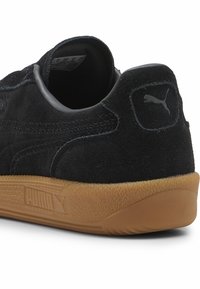 Svarta mockasneakers med en texturerad svart gummisula, med en grå Puma-logotyp på hälen och enkel sömdekoration på sidorna.