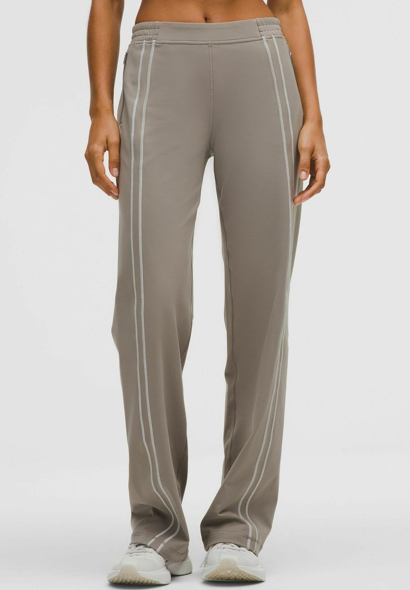 Világos barna sportnadrág, rugalmas derékkal, függőleges fehér csíkokkal és lazított fazonnal. Simább, nyújtható anyagból készült.