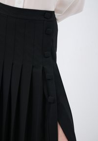 Gonna nera a pieghe realizzata in tessuto con bottoni laterali e uno spacco aperto. La texture è liscia e il design presenta pieghe nette.