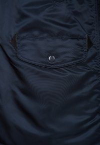 Interno della giacca blu navy con una tasca a bottone automatico, dettagli in tessuto nero e una texture liscia e lucida. Cuciture visibili lungo i bordi.