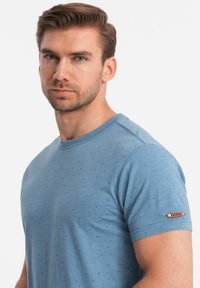 Camiseta de manga corta azul claro con pequeños puntos oscuros, escote redondo y un detalle metálico en la manga. Tejido suave con una superficie texturizada.