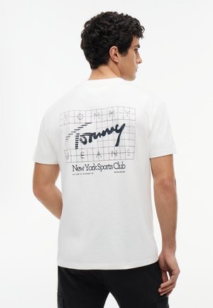 Mężczyzna tyłem, ubrany w białą koszulkę z czarną kratką i napisem „Tommy Jeans New York Sports Club” na plecach, czarne spodnie.