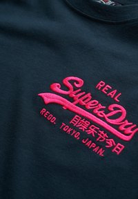 Camiseta de algodón negra con texto bordado en rosa que dice "SuperDry," "REAL," "REGD. TOKYO, JAPAN," con una textura suave.