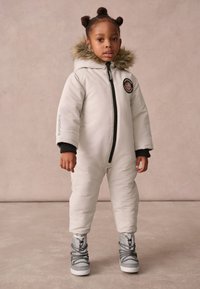 Padded white snowsuit with a black zipper and hood, featuring fur trim and a round patch. Gray boots with lace detailing complete the outfit.
