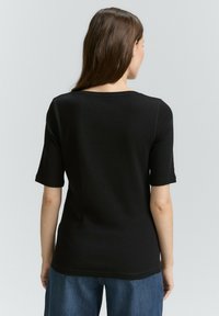 Černý top s krátkým rukávem a kulatým výstřihem vyrobený z hladké tkaniny. Má volný střih a jednoduchý design, zobrazený ze zadního pohledu.