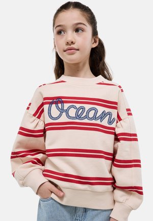 Fille aux cheveux foncés portant un sweat crème avec des rayures rouges et "Ocean" brodé en bleu, mains dans les poches d'un pantalon à rayures bleu clair.