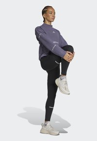 Camiseta atlética de manga larga morada con tejido texturizado y detalles reflectantes, combinada con leggins negros y zapatillas de running gris claro.