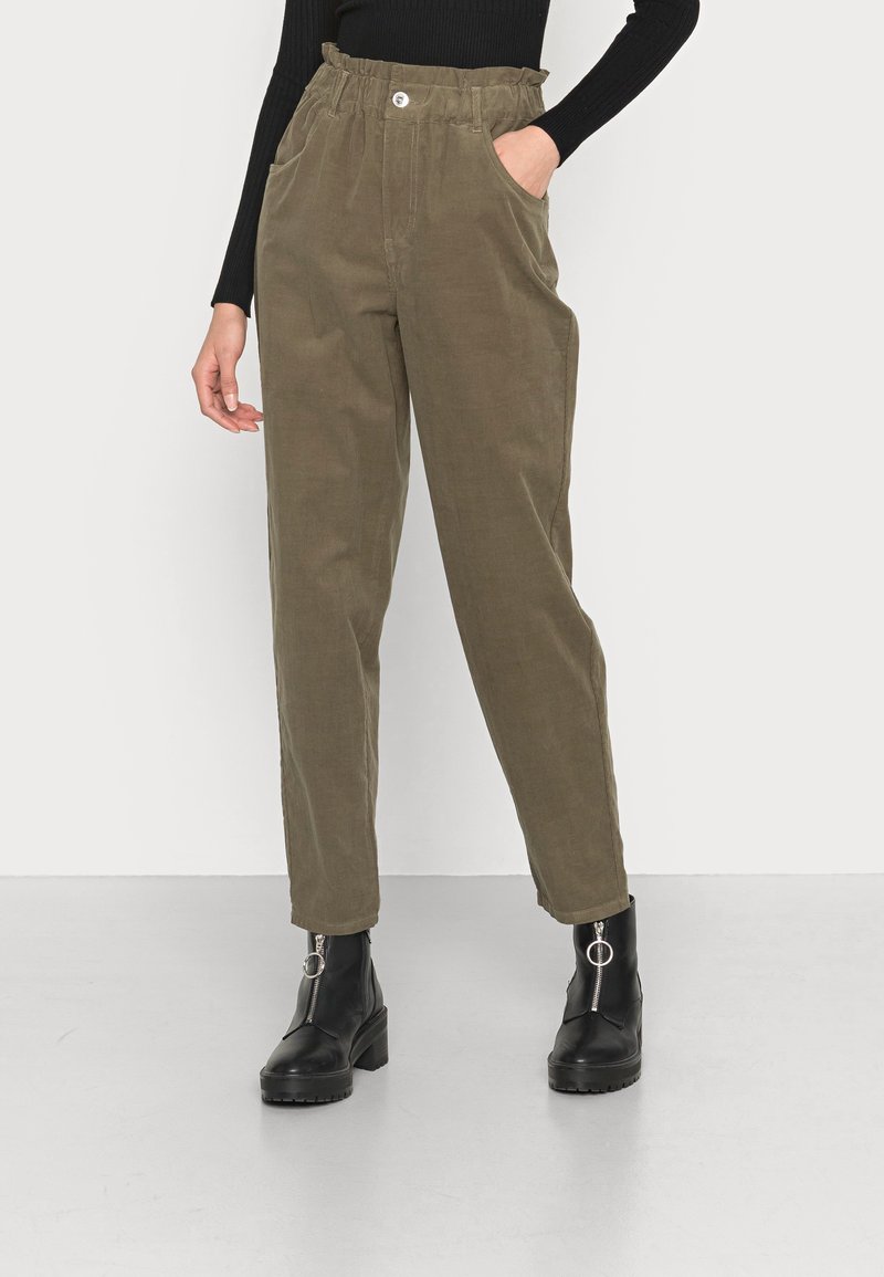 Pantalons en velours côtelé vert olive avec taille haute et ceinture élastique. Poches avant et fermeture à bouton unique. Associés à des bottines noires.