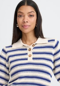 Pletena prugasta polo majica v kremni in mornarski barvi, z teksturirano zasnovo, tremi velikimi gumbi in klasičnim ovratnikom.