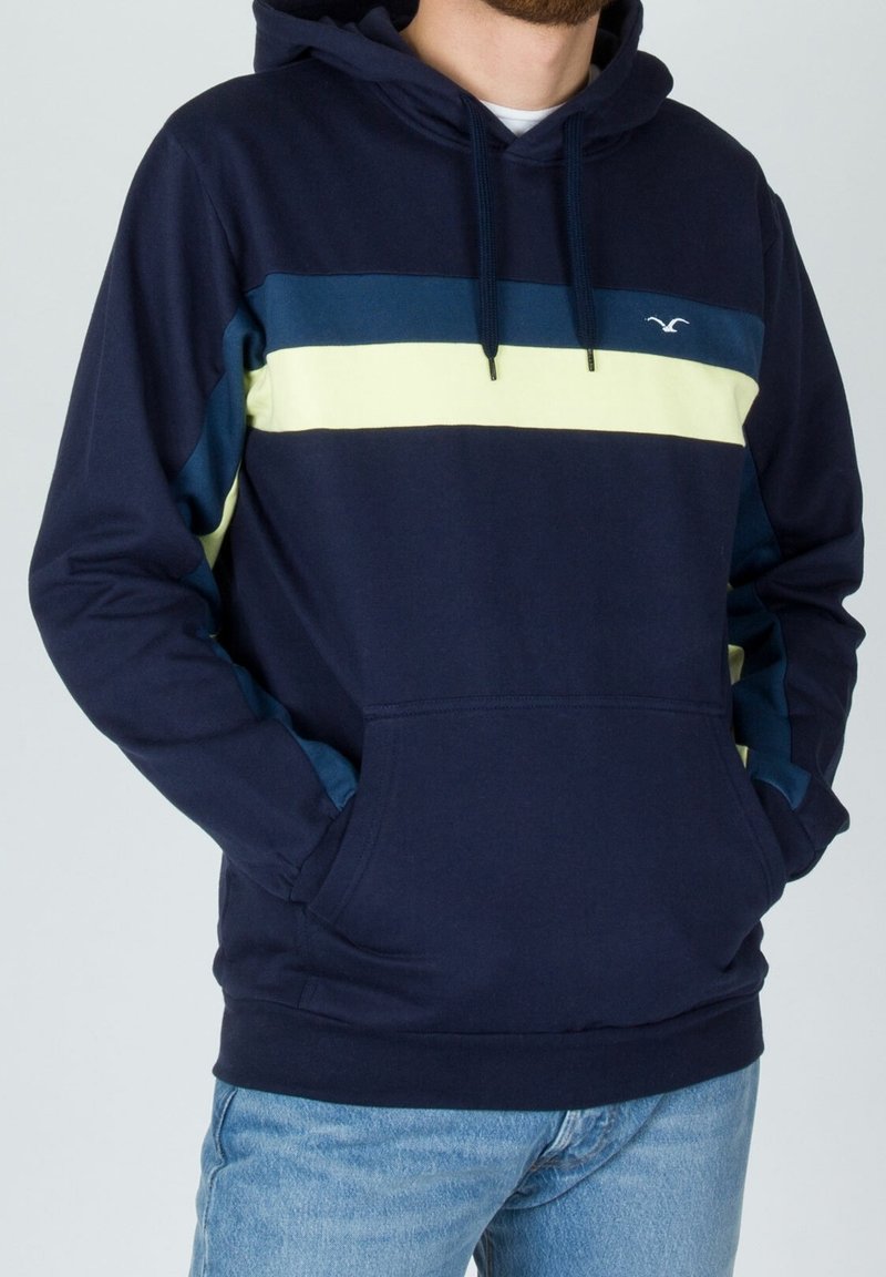Uomo che indossa un hoodie blu navy con strisce orizzontali blu e giallo pallido e jeans azzurri chiari, con le mani nella tasca frontale dell'hoodie.
