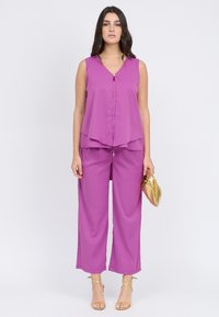 Top viola senza maniche con scollo a V e dettaglio di bottoni, abbinato a pantaloni viola a gamba larga. La modella tiene una clutch dorata. Finitura in tessuto liscio.