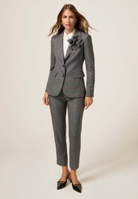 Vrouw in een grijs geblokt maatpak met een grote stoffen bloem op de revers, een witte blouse en zwarte puntige hakken, staand tegen een effen achtergrond.