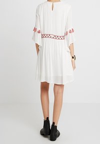 Vestido blanco con mangas cortas acampanadas, que presenta borlas rosas y bordados decorativos en la cintura. La parte de atrás tiene una abertura en forma de lágrima. Botas negras hasta el tobillo.