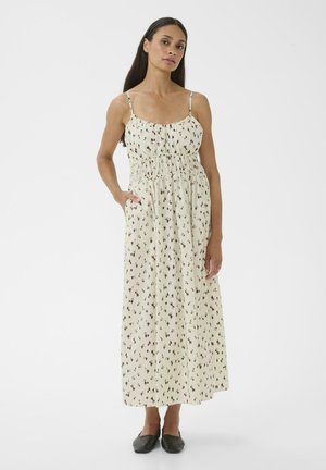 Frau trägt ein cremefarbenes Maxikleid mit dünnen Trägern und kleinem dunklem Blumenmuster sowie schwarze flache Schuhe, steht mit einer Hand in der Tasche.
