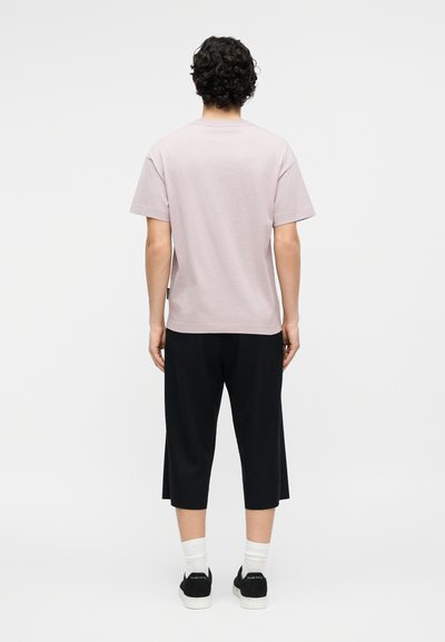 Άτομο με σγουρά σκουρόχρωμα μαλλιά, που στέκεται με την πλάτη προς την κάμερα, φοράει ανοιχτόχρωμο κοντομάνικο πουκάμισο, μαύρο κοντό παντελόνι, λευκές κάλτσες και μαύρα αθλητικά παπούτσια.