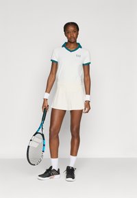 Λευκό πόλο μπλουζάκι τένις με τόνους πετρόλ στο γιακά, συνδυασμένο με φούστα σε κρεμ απόχρωση. Μαύρα παπούτσια τένις, κρατώντας ρακέτα. Απλός και σπορ σχεδιασμός.