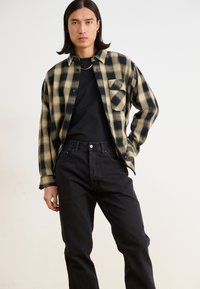 Svart og brun rutet flanellskjorte med krage og enkelt brystlomme, brukt over en svart T-skjorte og kombinert med svarte jeans.