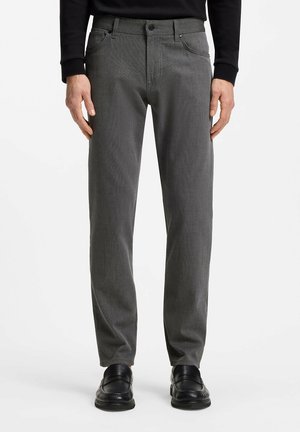 Hombre vistiendo pantalones grises de corte recto con bolsillos delanteros y zapatos de vestir negros, de pie contra un fondo blanco liso.