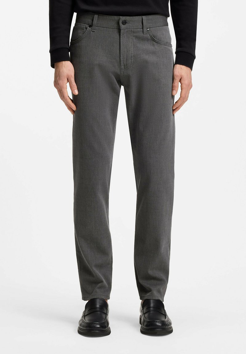 Homme portant un pantalon gris à coupe droite avec des poches avant et des chaussures habillées noires, debout devant un fond blanc uni.