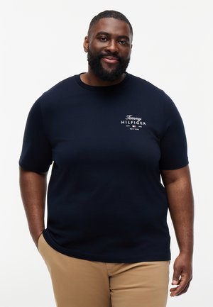 Usměvavý vousatý muž v tmavě modrém tričku Tommy Hilfiger a béžových kalhotách, stojící s jednou rukou v kapse na jednobarevném pozadí.