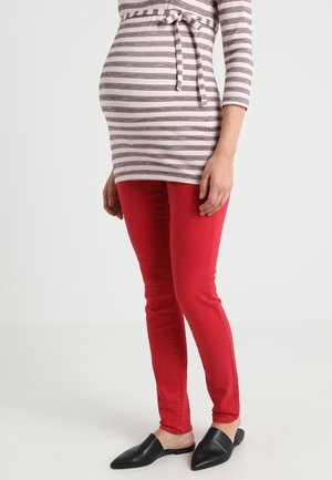 Femme enceinte portant un haut à manches longues rayé avec un détail de nouage, un pantalon rouge et des chaussures noires faciles à enfiler, debout sur un fond uni.