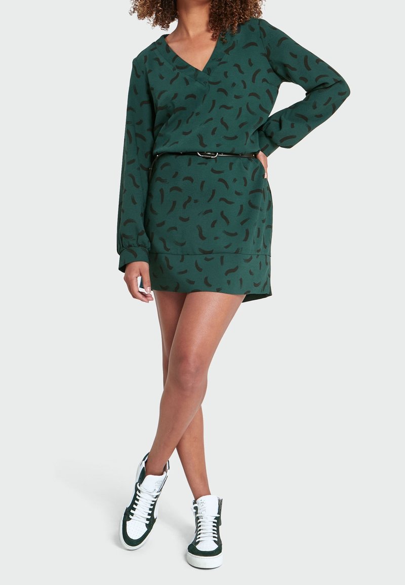 Mujer con un mini vestido de manga larga verde con patrones abstractos negros, cinturón negro y zapatillas altas en blanco y negro, posando con la mano en la cadera.