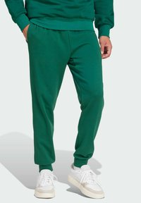 Pantalones de chándal verdes hechos de tela suave, con una cintura y puños elásticos. Combinados con zapatillas blancas. Textura suave, diseño ajustado.