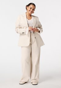 Vrouw die glimlacht, gekleed in een beige pak met kantachtige manchetten, een witte kanttop, gouden sieraden en wit met studs schoenen, staand voor een effen witte achtergrond.