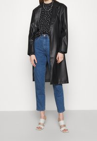Long cappotto in pelle sintetica nera, blusa a pois, jeans blu a vita alta e mule grigi chiari con tacco e cinturini incrociati.