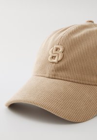 Beige kordfløyelscaps med en buet kviste, som har en brodert logo på forsiden; teksturen er ribbet, noe som gir en myk følelse.