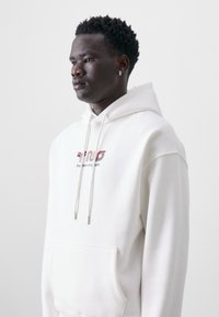 Hvit hettegenser laget av mykt stoff, med en logo i rødt og grønt, justerbare snorer og en frontlomme.